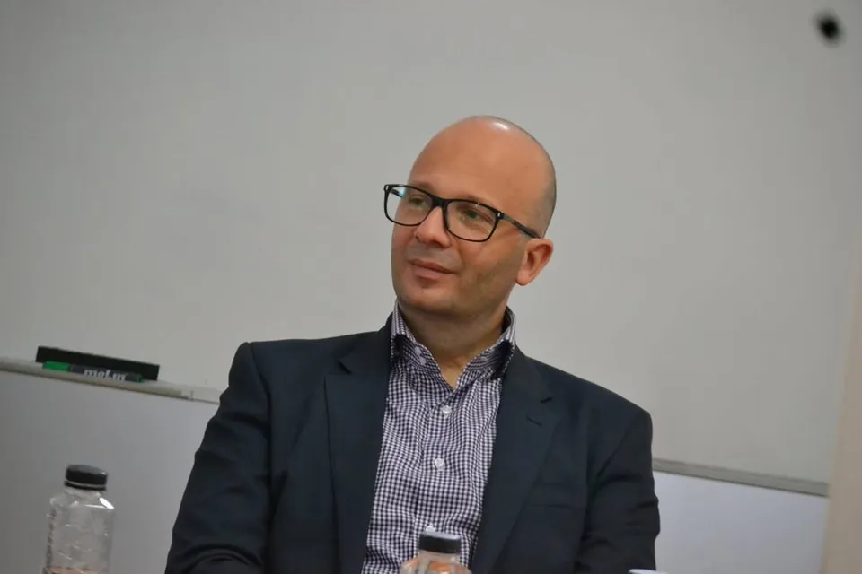 Un bărbat cu ochelari, purtând sacou închis și cămașă închisă la culoare, stă așezat în fața unui fundal alb simplu.