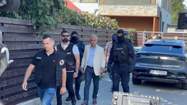 Un grup de bărbați merge pe un trotuar lângă o clôtură de lemn și o mașină neagră, unii purtând veste negre și măști.