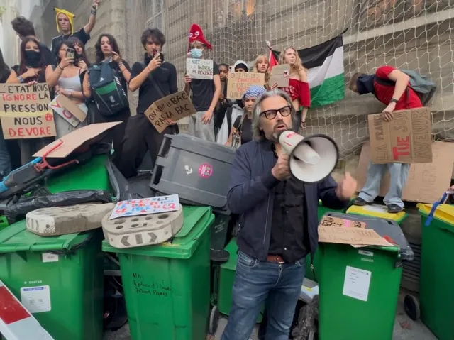 Un bărbat cu ochelari și un megafon stă în prim-plan lângă containere verzi de gunoi, în timp ce un grup de persoane protestează în spate.