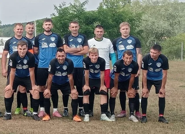 O echipă de nouă bărbați în echipamente sportive albastre și negre pozează într-o poziție de grup pe un teren de fotbal cu iarbă uscată.