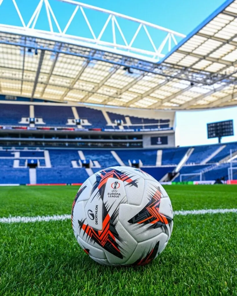 Un fotbal alb cu modele geometrice negre și roșii este plasat pe un teren de gazon verde cu o linie albă.