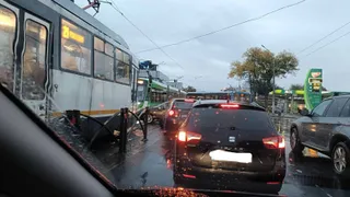 Un tramvai gri și un autoturism negru sunt vizibile pe un carosabil umed sub un cer înnorat.