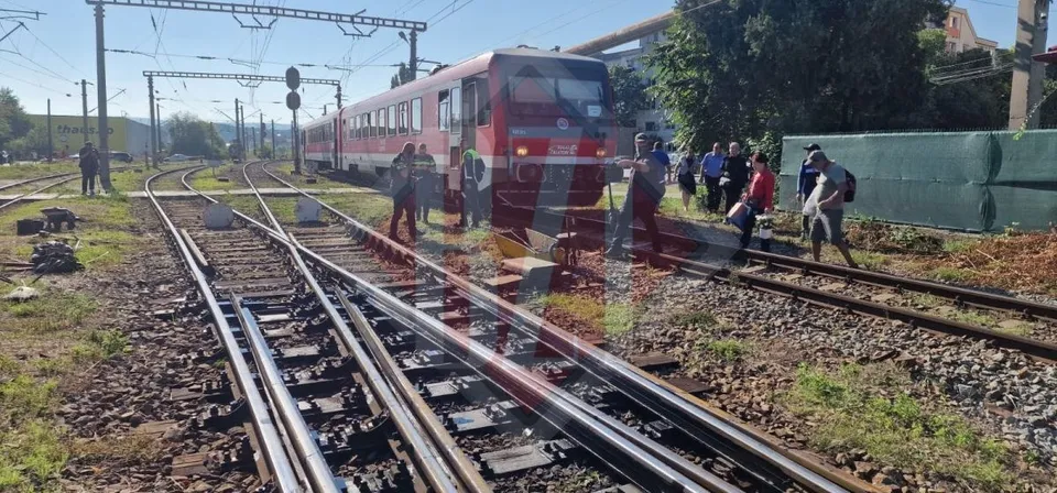 Un tren roșu stă pe șine feroviare lângă un grup de persoane care stau pe balastul de pietriș.
