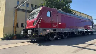 O locomotivă electrică de culoare roșie cu detalii gri este oprită pe o cale ferată lângă un clădire galbenă sub cerul senin.