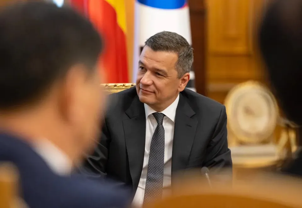 Un bărbat cu părul scurt și costum negru stă la o masă, privind spre o persoană aflată în prim-plan, desfocată.