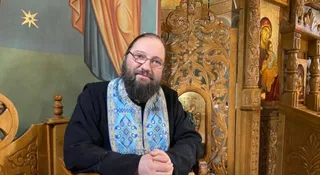Un bărbat cu barbă și ochelari poartă o vestă albastră decorată și stă în fața unui obiect din lemn sculptat lângă un iconostas.