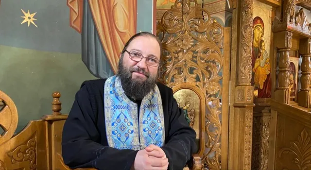 Un bărbat cu barbă și ochelari poartă o vestă albastră decorată și stă în fața unui obiect din lemn sculptat lângă un iconostas.