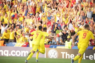 Câțiva jucători de fotbal în echipamente galbene celebrează pe un teren verde în fața unei mulțimi de spectatori care poartă haine galbene și roșii.