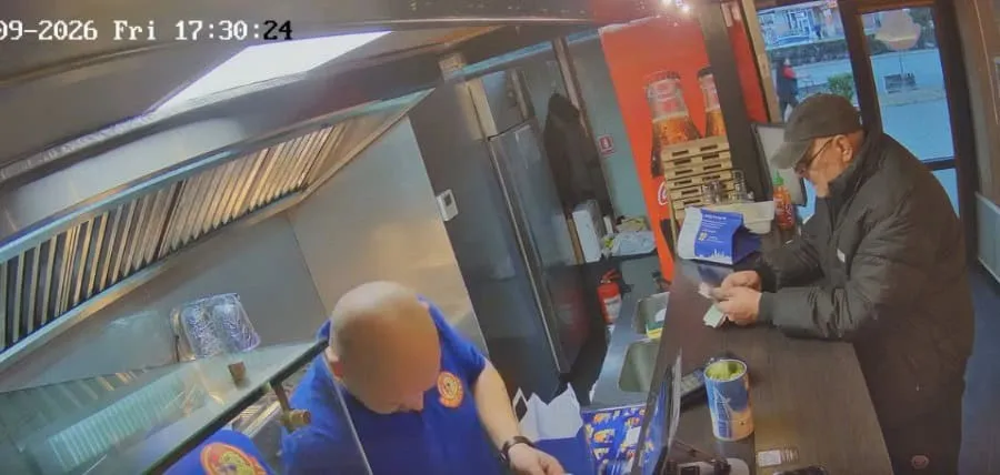 Un bărbat cu o șapcă și o jachetă neagră stă la un tejghea lângă un angajat cu tricou albastru într-un spațiu interior.