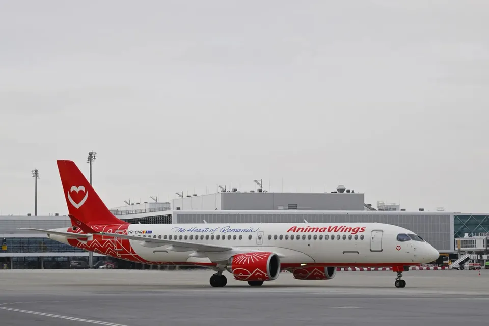 Un avion alb cu accente roșii și inscripția AnimaWings stă pe o pistă de aeroport sub un cer înnorat.