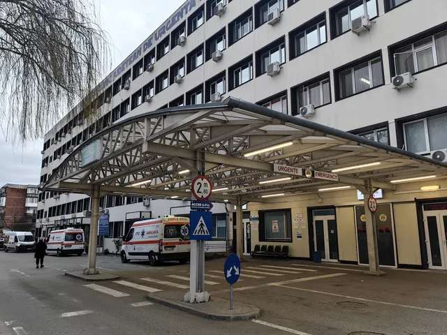 O clădire albă cu ferestre multiple este lângă o structură metală acoperită care adăpostește două ambulanțe albe parcate pe un teren asfaltat.
