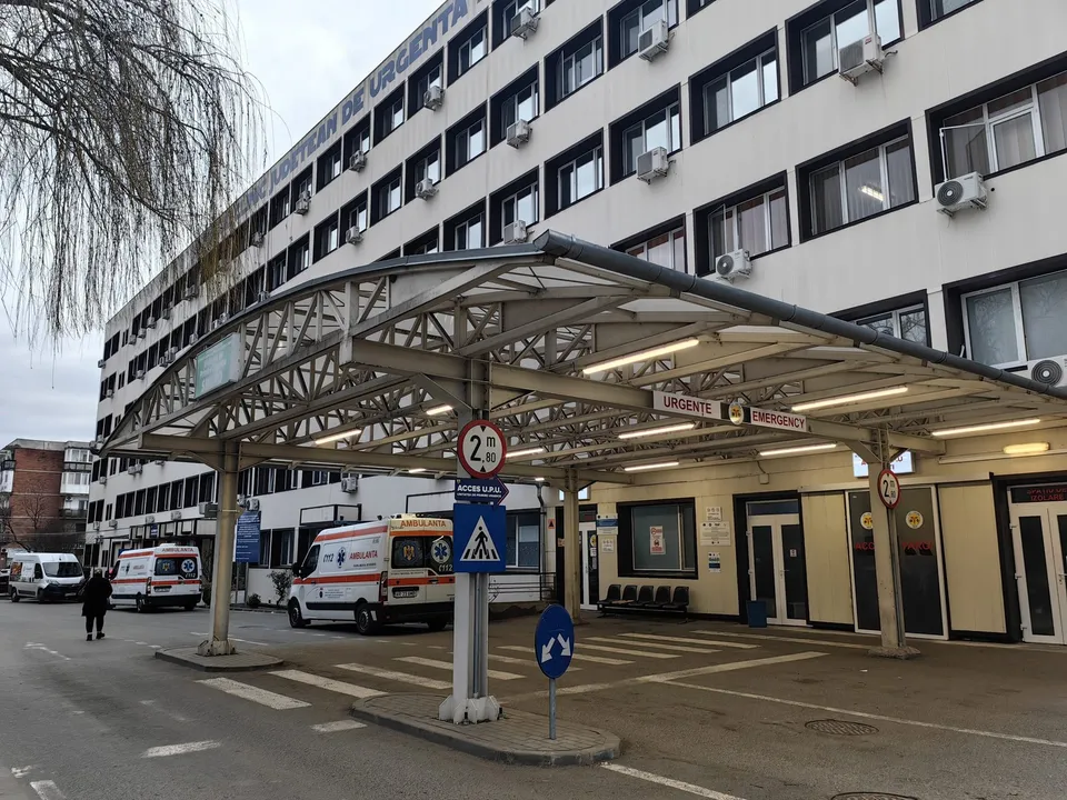 O clădire albă cu ferestre multiple este lângă o structură metală acoperită care adăpostește două ambulanțe albe parcate pe un teren asfaltat.