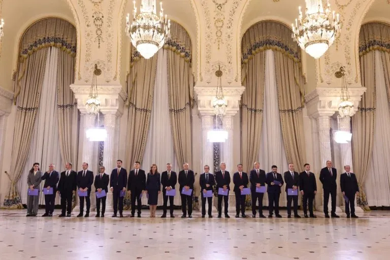 Un grup de persoane îmbrăcate în costume oficiale stă în rând într-o sală încăpătoare cu candelabre mari și draperii înalte.