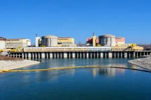 O clădire industrială cu două structuri cilindrice gri se află lângă un corp de apă sub un cer senin.
