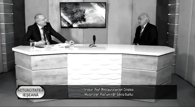 Un bărbat cu ochelari și costum stă la o masă de studio lângă un alt bărbat cu părul alb și costum într-un cadru de televiziune.