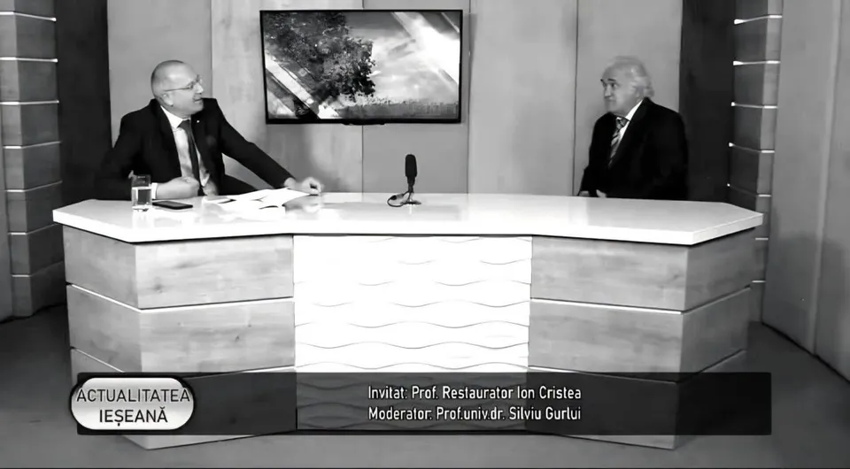 Un bărbat cu ochelari și costum stă la o masă de studio lângă un alt bărbat cu părul alb și costum într-un cadru de televiziune.