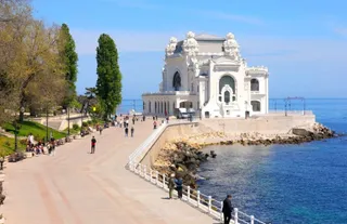 O clădire albă de arhitectură monumentală se află pe malul mării, lângă un drum pietonal pe unde circulă oameni sub un cer senin.