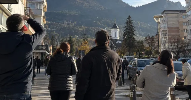 Cinci persoane stau cu spatele, privind spre un deal împădurit cu clădiri și un turn alb sub un cer senin.