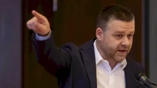 Un bărbat cu barbă, purtând sacou închis și cămașă albă, stă în fața unui microfon și arată cu degetul spre stânga.