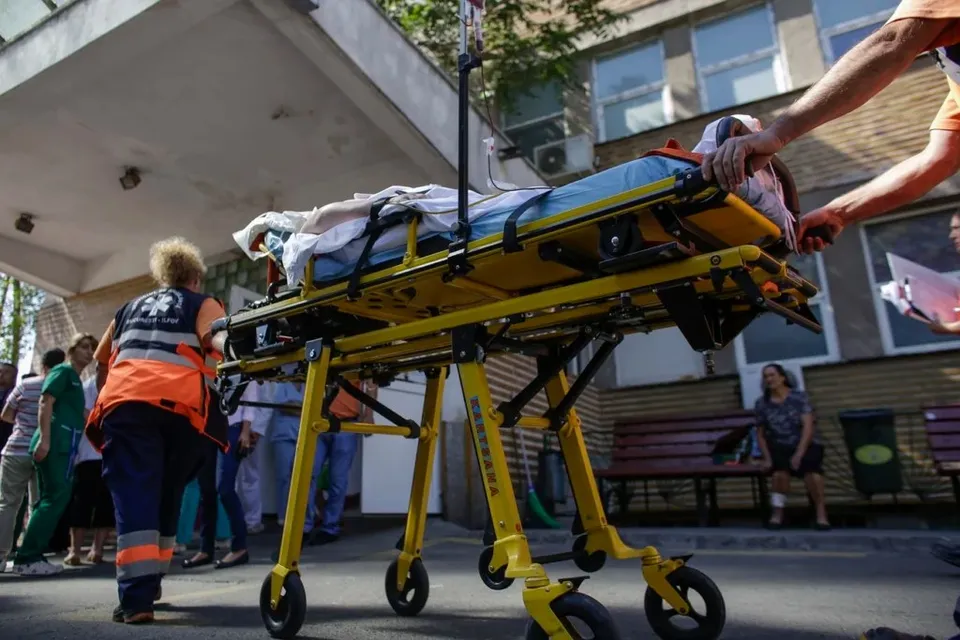 O persoană este transportată pe o targă medicală galbenă, împinsă de un paramedic, în fața unei clădiri.