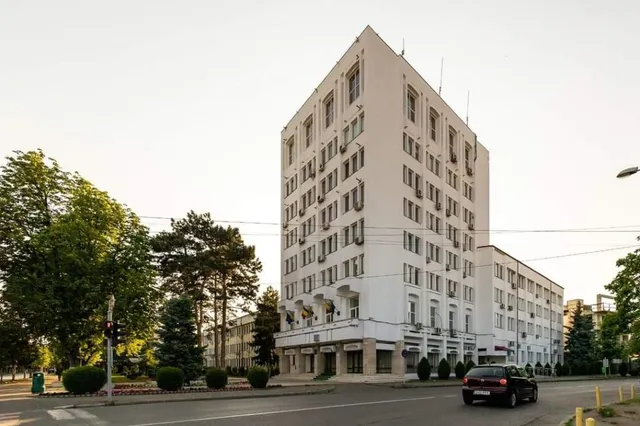 O clădire albă de mai multe etaje cu ferestre rectangulare se ridică lângă un drum cu un automobil negru și copaci verzi.