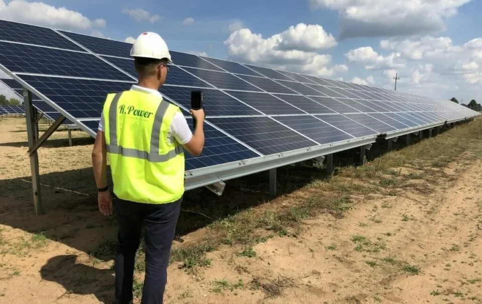 Un bărbat cu cască albă și vestă reflectorizantă verde se uită spre rânduri de panouri solare instalate pe un teren de pământ uscat sub un cer înnorat.