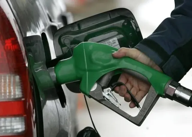 O mână ține un pistol de pompat combustibil verde introdus în orificiul de umplere al unui autovehicul gri.