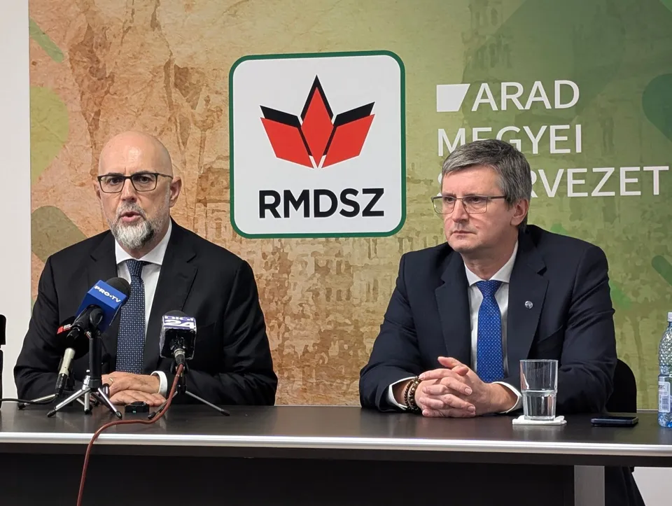 Doi bărbați în costume și cravate stau la o masă în fața unui panou cu un logo și textul RMDSZ.