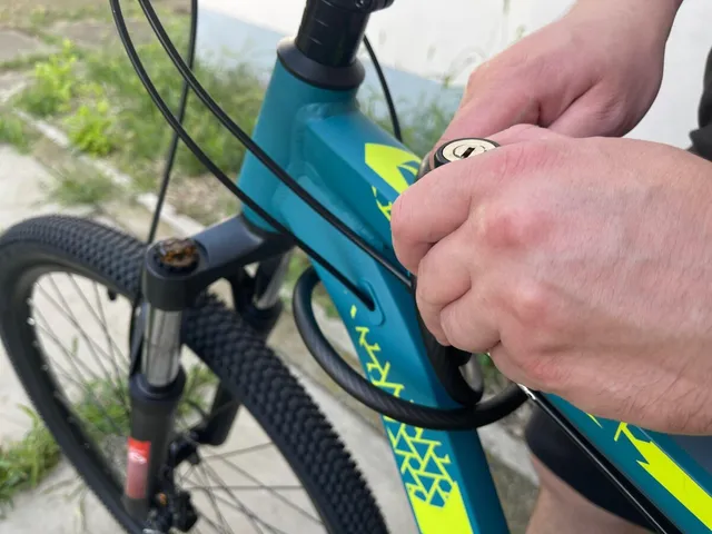 Mâinile unui om folosesc o cheie pentru a manipula un lacăt de tip cablu în jurul cadrului unei biciclete de culoare albastră.