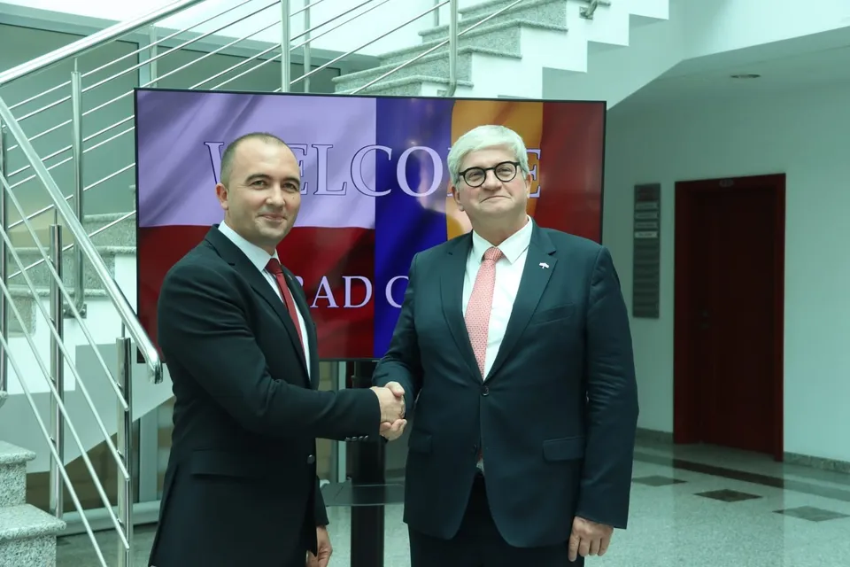 Doi bărbați în costume stau în picioare și se strâng în mână în fața unui panou colorat într-un interior cu scări metalice.