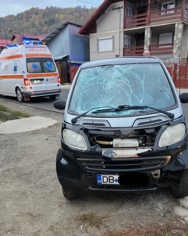O mașină gri cu parbrizul spart și partea din față deteriorată este parcată pe un drum, cu o ambulanță albă și roșie vizibilă în fundal.