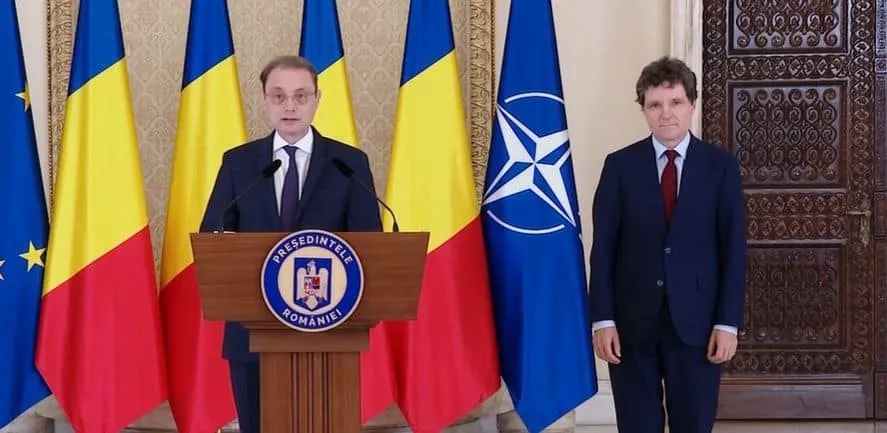 Un bărbat stă în spatele unui podium cu emblema României, iar lângă el un alt bărbat stă în picioare în fața unor steaguri.