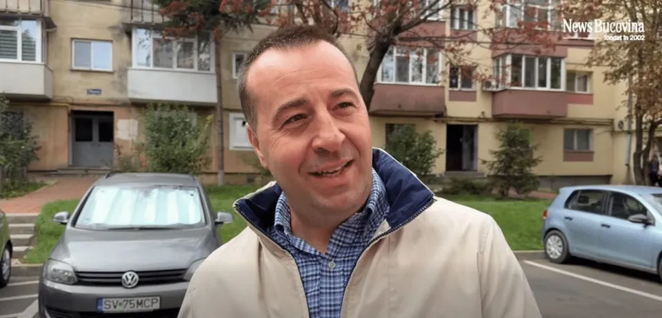 Un bărbat zâmbind poartă o cămașă în carouri și o jachetă deschisă, având în fundal clădiri de locuințe și mașini parcate.