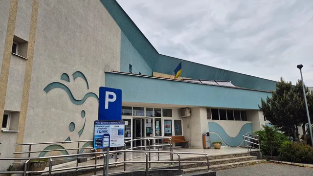 O clădire cu fațadă gri și albastră prezintă un semn de parcare albastru și o rampă metalică în prim-plan.