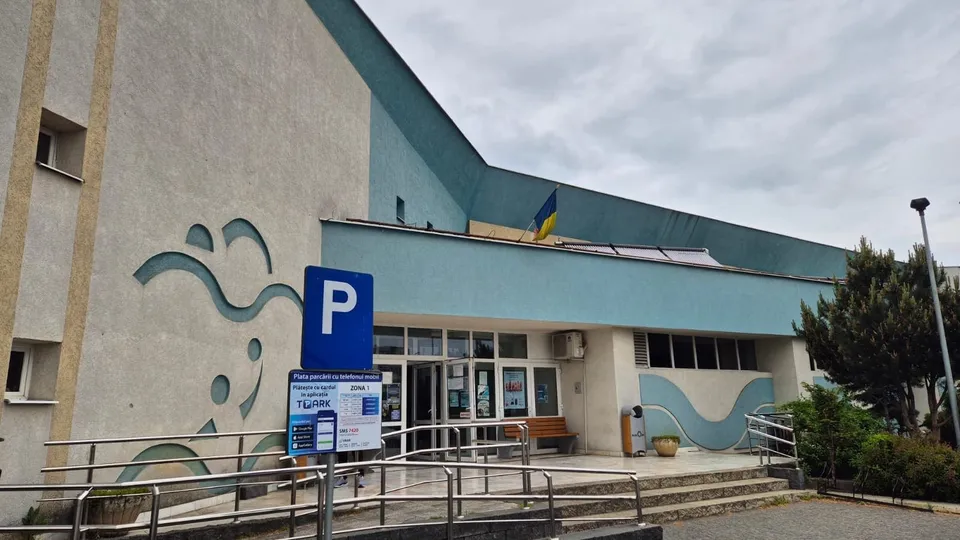 O clădire cu fațadă gri și albastră prezintă un semn de parcare albastru și o rampă metalică în prim-plan.