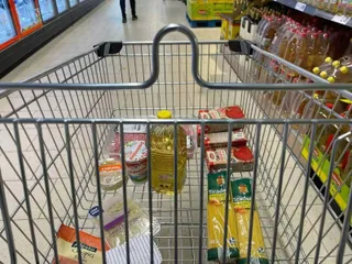 Un coș de cumpărături metalic conține sticle de ulei, cutii de alimente și pungi de produse într-un magazin.