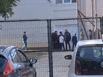 Un grup de persoane stă adunat în spatele unui gard metalic, lângă un vehicul alb și o mașină neagră.