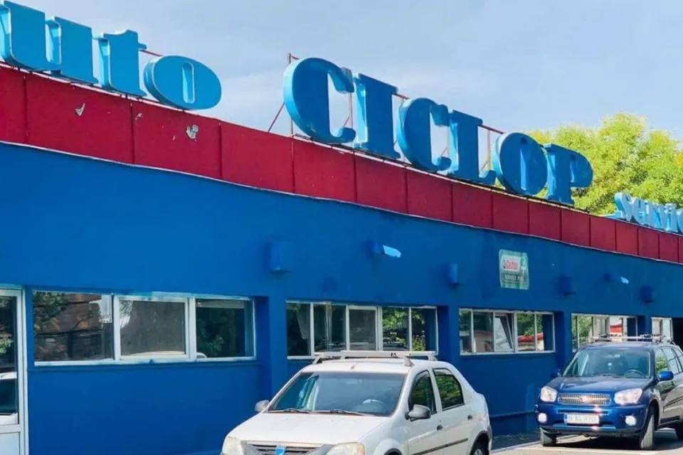 O clădire albastră cu un semn mare albastru pe acoperiș stă lângă două mașini parcate pe un drum.