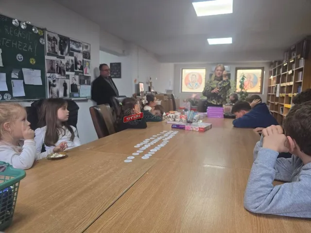 Copii și adulți stau la o masă lungă de lemn într-o încăpere cu rafturi de cărți și afișe pe perete.