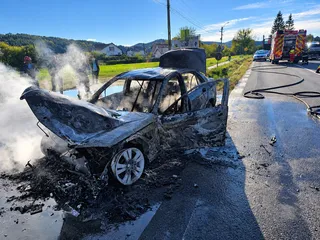 Un autoturism carbonizat stă pe marginea unui drum asfaltat lângă un nor de fum alb, sub un cer senin.