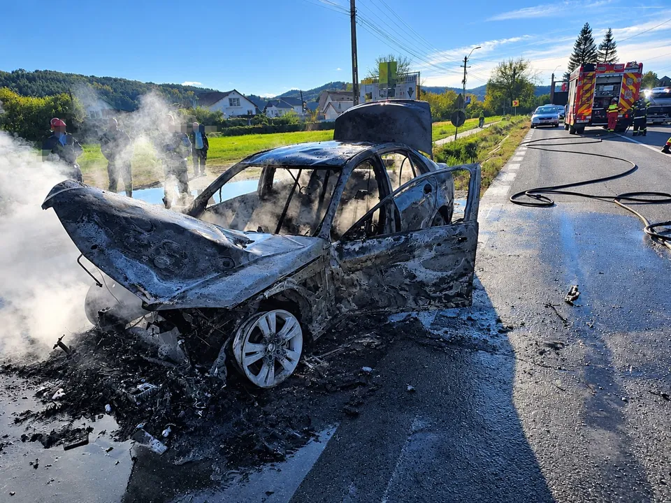 Un autoturism carbonizat stă pe marginea unui drum asfaltat lângă un nor de fum alb, sub un cer senin.