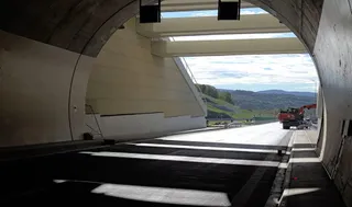 Interiorul unui tunel de beton cu o porțiune de asfalt întins și un utilaj de construcții vizibil la ieșirea spre un peisaj colinar.