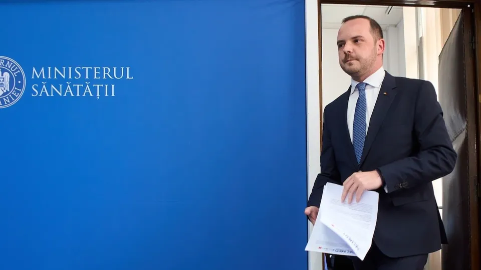 Un bărbat în costum bleumarin, cravată albastră și cămașă albă, merge în picioare ținând hârti albe în mână lângă un panou albastru cu textul Ministerul Sănătății.