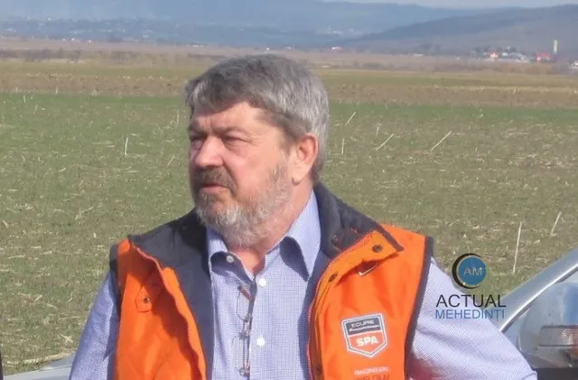 Un bărbat cu barbă și părul cărunt poartă o vestă portocalie peste o cămașă albastră, stând în fața unui câmp deschis.