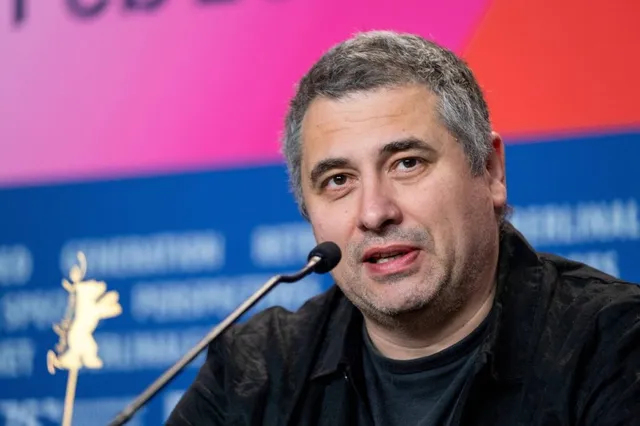 Un bărbat cu părul scurt și cămășă neagră vorbește în fața unui microfon pe un fundal colorat cu text și un simbol auriu.