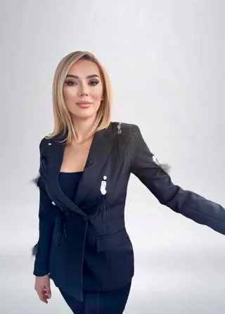 O femeie cu părul blond și bluză neagră poartă un sacou negru decorat cu pene și elemente albe pe un fundal gri deschis.