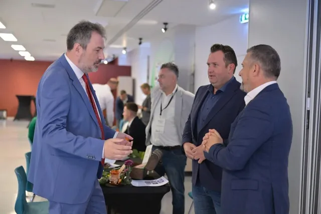 Un grup de bărbați în haine office stau în picioare și discută într-o încăpere luminoasă cu pereți albi și un perete roșu în fundal.
