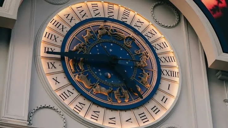 Un ceas circular mare cu cifre romane pe un fundal alb și un cadran central albastru decorat cu simboluri ale zodicilor astrologice și stele.