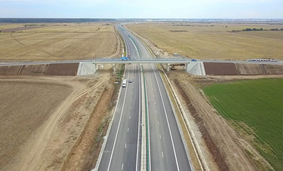 O autostradă cu mai multe benzi traversează un câmp deschis, fiind traversată de un pod de o structură gri sub un cer înnorat.