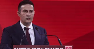 Un bărbat cu părul scurt, purtând costum închis la culoare, cămașă albă și cravată închisă, vorbește la un podium cu microfoane în fața unui fundal roșu.